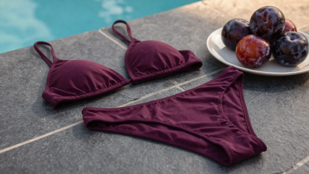 Deep plum purple bikini beside ripe plums on dark slate, showing the rich shade, fabric sheen and fit